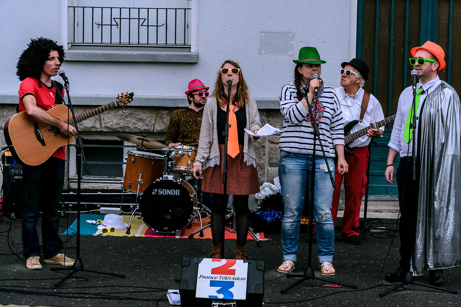 Six eccentrically dressed musicians playing on a Paris street, behind a banner for a French television station. 