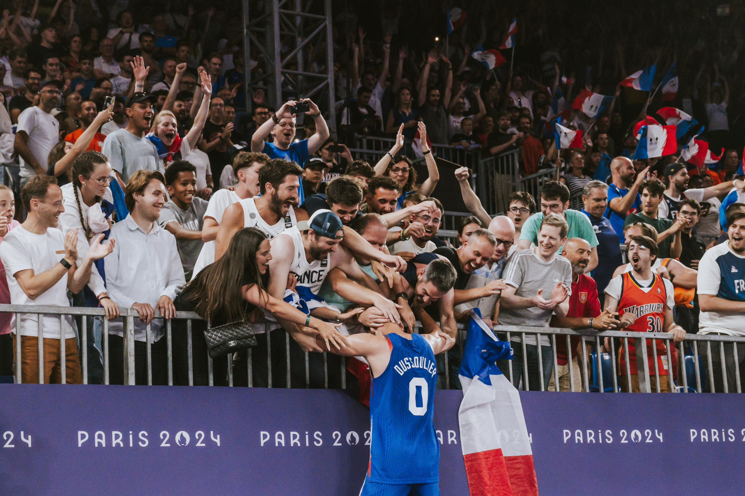 Olympic fans at the Paris 2024 reaching over the guardrails to embrace French basketballer Dussoulier. A French flag is draped over the guardrails to the right of Dussoulier. 