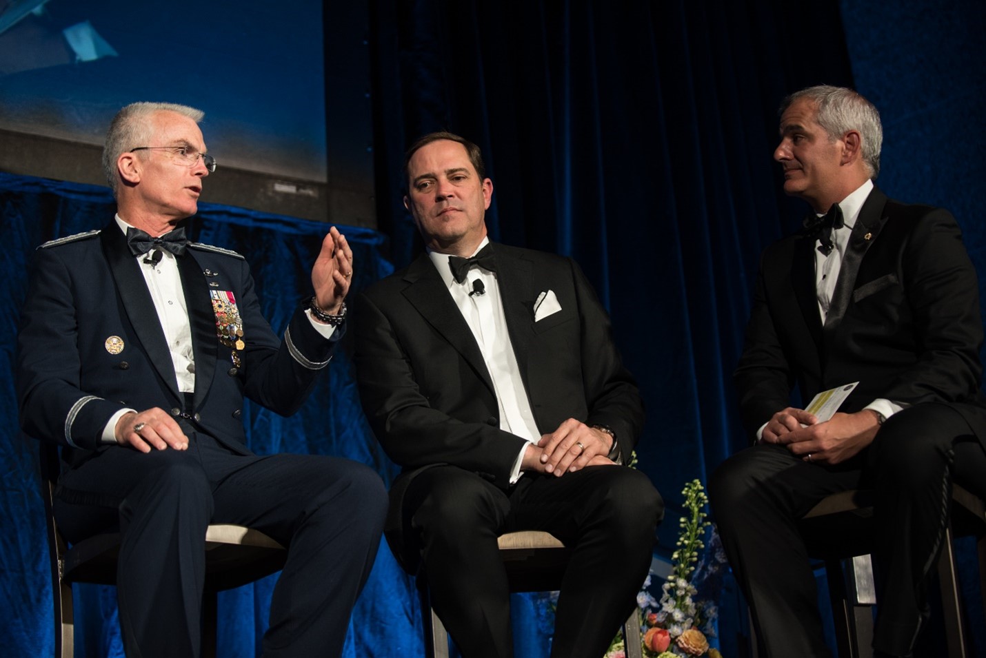 CEO Chuck Robbins dressed in formal suit and tie, seated on what looks like a stage in front of a projector screen between two similarly attired middle-aged white men. 