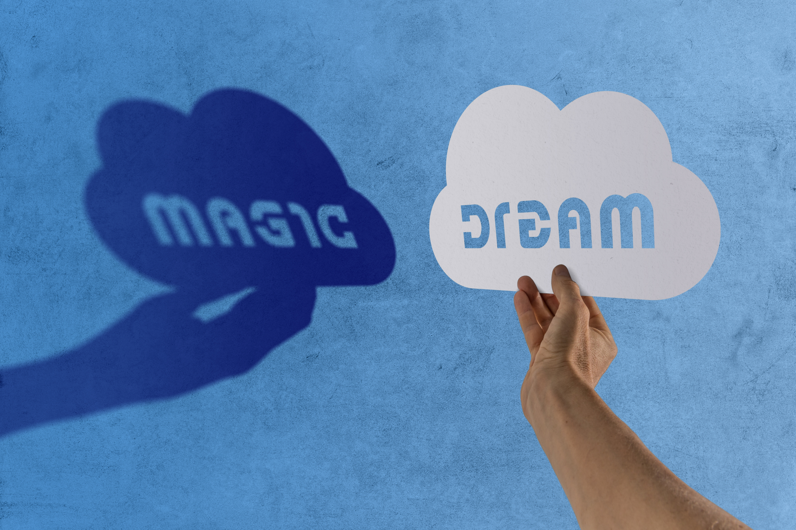 A metaphor for AI sleight of hand: Paper cut-out of a cloud with the word "dream" cut out held in someone's hand that reads "magic" when its reflection is projected against the wall.