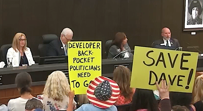 Photograph of city council meeting attended by the public holding up handwritten signs reading "Save Dave!" among others