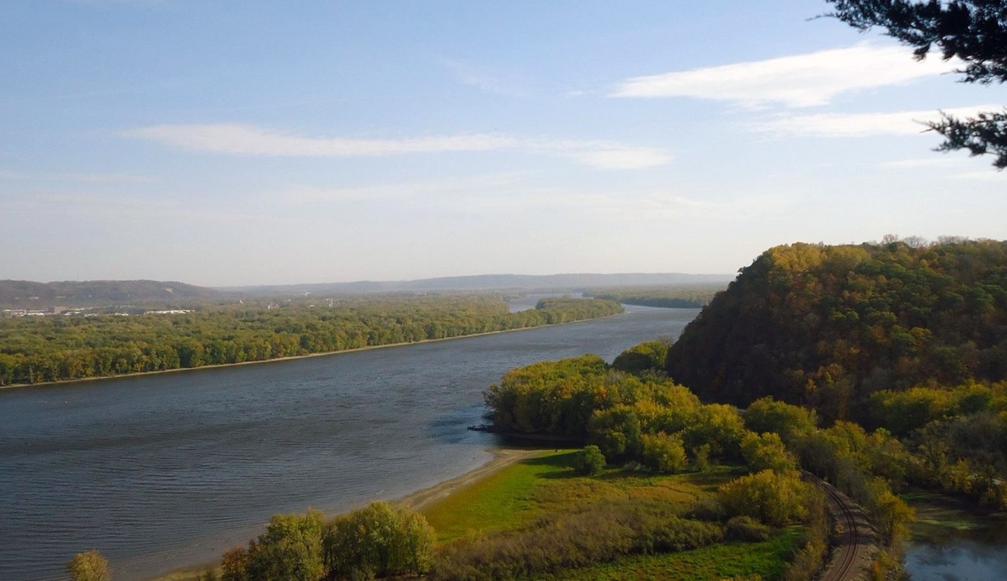 Photograph of lush green bluffs along a sediment-heavy river, across from forested planes