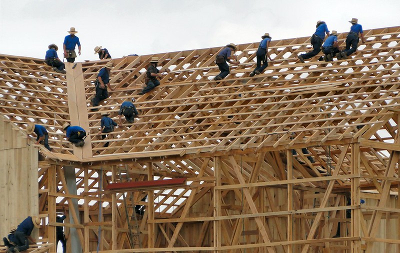 Over a dozen Amish men stand, working, on the partially-constructed roof of a wooden barn.