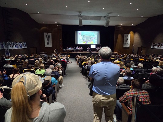 a large auditorium full of people facing a stage where several people sit at a table in front of a screen displaying a map
