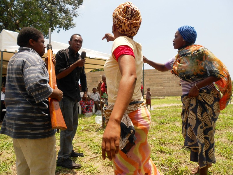 Four people are engaged in a performance. Two angry people point at a third person who holds a microphone and looks defensive. A fourth person holds a rifle.