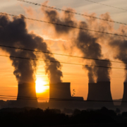 Giant plumes of smoke rise from four smokestacks as the sun sets in the background, with power lines in the foreground.
