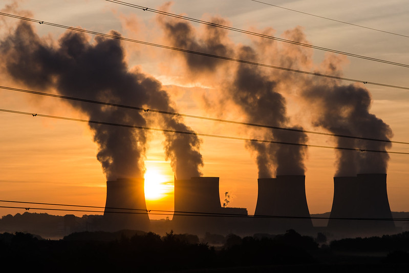 Giant plumes of smoke rise from four smokestacks as the sun sets in the background, with power lines in the foreground.