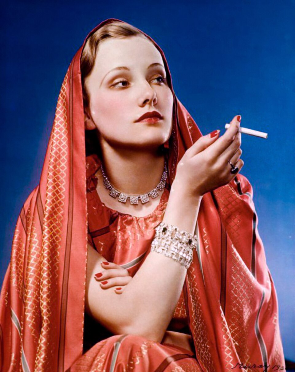 A glamorously-dressed woman holds a cigarette between her fingers and gazes to the side.