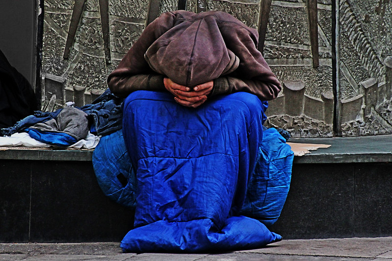 A person sits outside on a stair with their legs in a sleeping bag and their head in their hands.