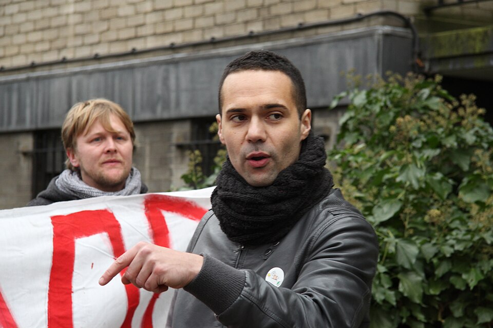 Bastani points, his mouth open in speech, and a man stands behind him holding a white sign with big red lettering.