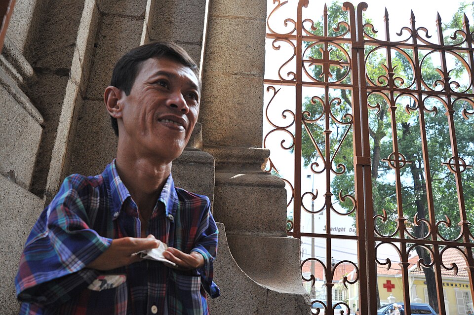 A person in a plaid shirt with short arms and two to three fingers per hand stands in front of a gate.