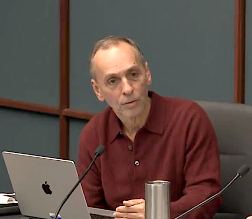 man sits in front of a laptop at a desk and earnestly speaks into a microphone