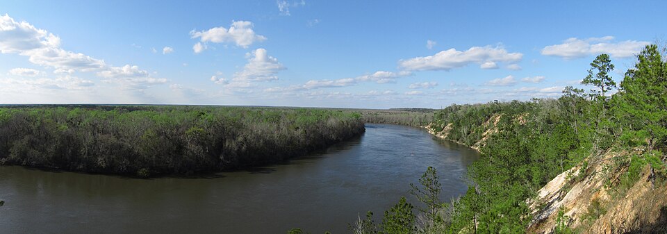 A river winds around a bend under a blue sky, with green shrubs on one side and bluffs on the other.