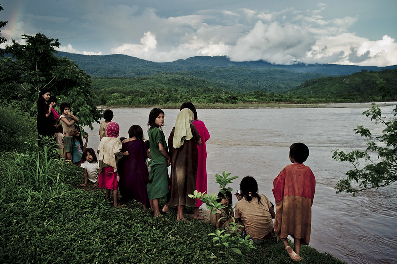 A group of children stand on a river bank, looking out at the river and lush, forested mountains beyond.