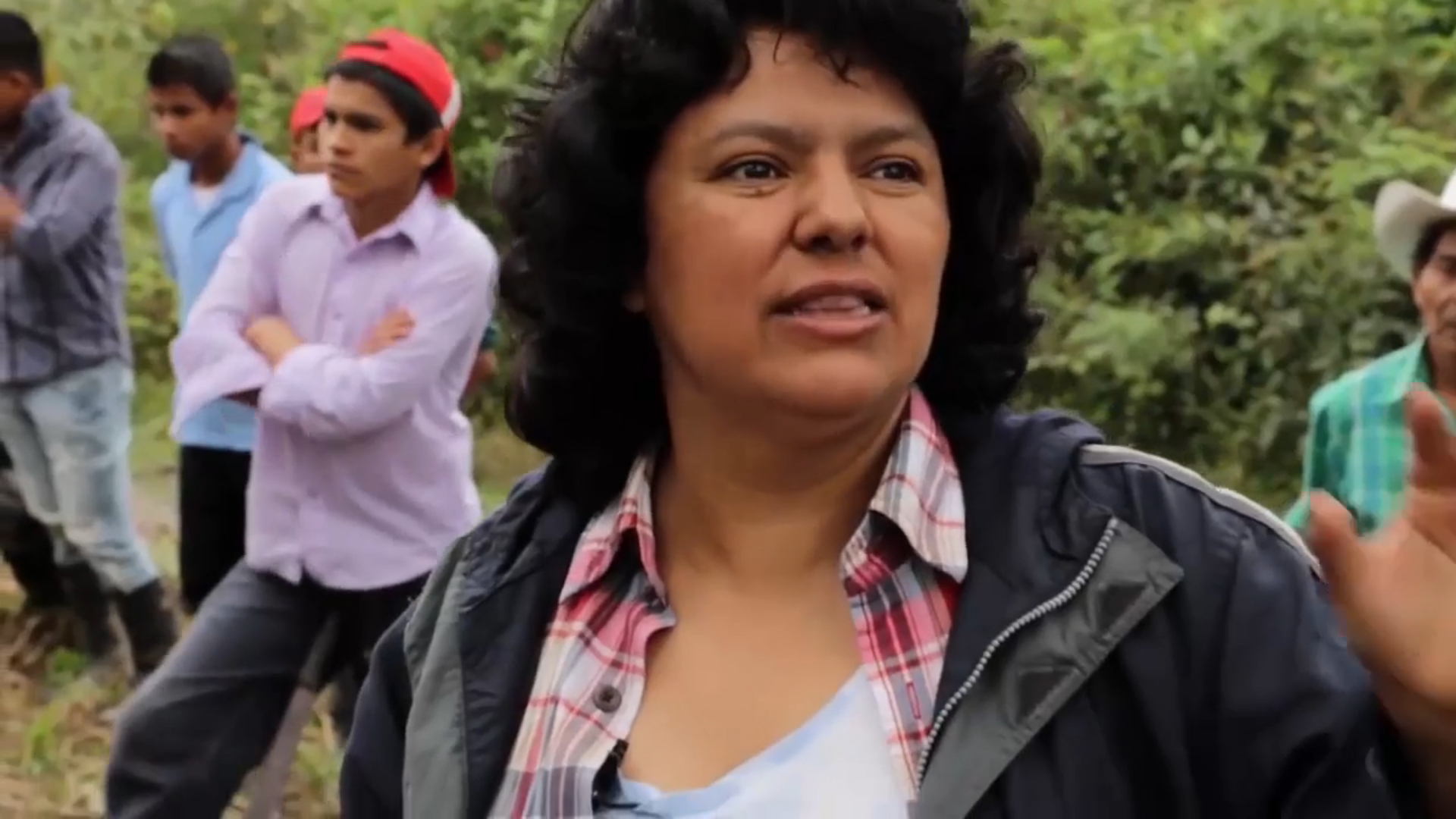 Candid shot of Berta Cáceres speaking with greenery and several people standing around in the background.