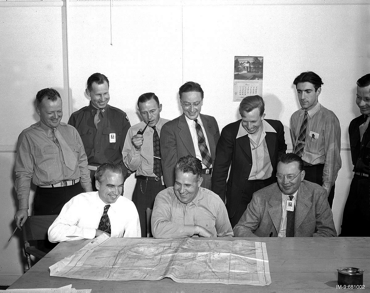 10 men gather around a table looking at a big map. All are smiling.