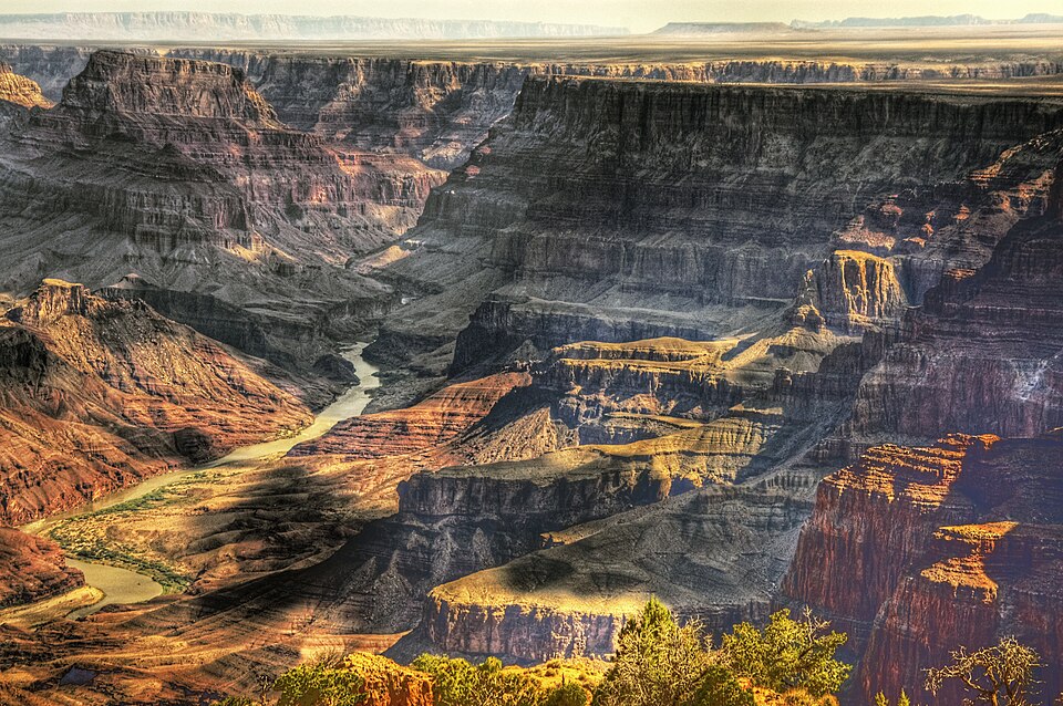 A wide-angle, aerial view of the Grand Canyon and landscape beyond. The canyon is drenched in golden light with a southwestern rainbow of red, orange, pink, and gray colors on the rocks. The Colorado River is seen in the base of the canyon.
