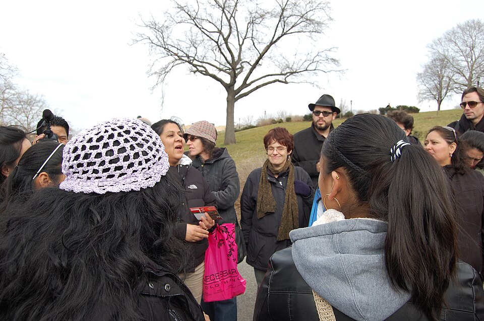 A diverse group of people stands outside wearing jackets, as one woman in the center of the group talks.