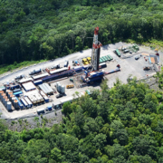 Aerial view of a frack pad. The pad is in a clearing surrounded by deciduous forest. You can make out the well itself, which looks similar to an oil derrick, a number of trucks parked on the sides of the access roads, and containers and tanks lined up on the opposite side of the pad from the well.