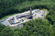Aerial view of a frack pad. The pad is in a clearing surrounded by deciduous forest. You can make out the well itself, which looks similar to an oil derrick, a number of trucks parked on the sides of the access roads, and containers and tanks lined up on the opposite side of the pad from the well.