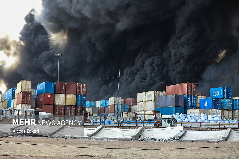 A giant plume of black smoke rises behind a yard full of shipping containers.
