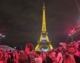 Picture of spectators, lit in red, standing in front of a brightly illuminated Eiffel Tower on a cloudy night