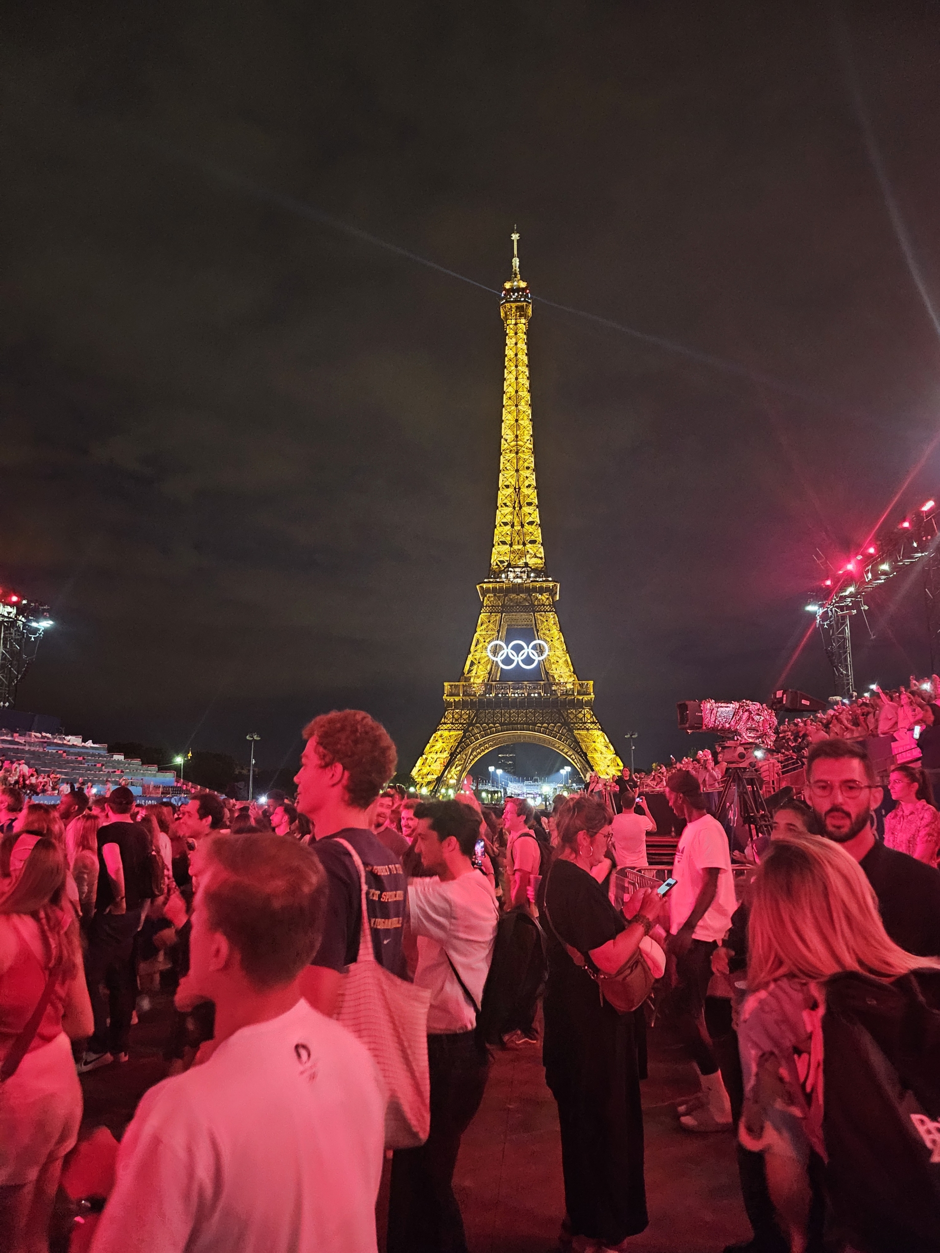 Picture of spectators, lit in red, standing in front of a brightly illuminated Eiffel Tower on a cloudy night