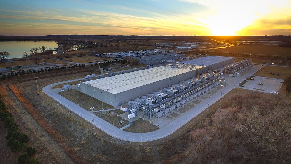 A large, warehouse-looking facility surrounded by fields and a body of water, with a sunset in the background.