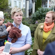 Housing_affordability_(34286937685) A woman with short blonde hair and an infant child in a knitted cap talks to a camera out of frame while another woman wearing a button listens.