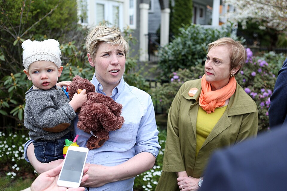 A woman with short blonde hair and an infant child in a knitted cap talks to a camera out of frame while another woman wearing a button listens.