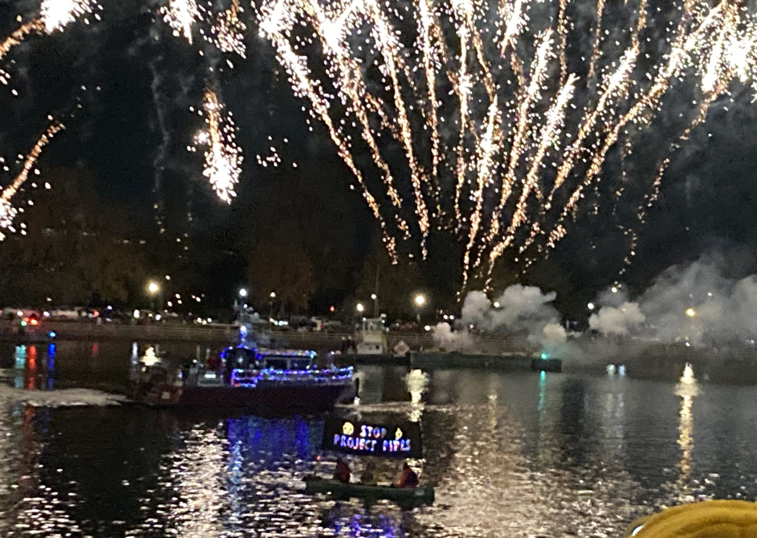 A canoe sits on the Potomac River at night in front of fireworks. Mounted to the canoe is a sign that says "STOP PROJECT PIPES" in Christmas lights.