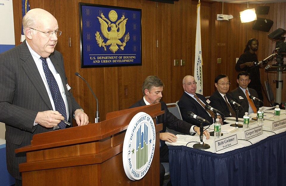 Close up of a speaker at the podium at U.S. Department of Housing and Urban Development headquarters to discuss the subprime mortgage crisis, with a row of panelists seated, listening.