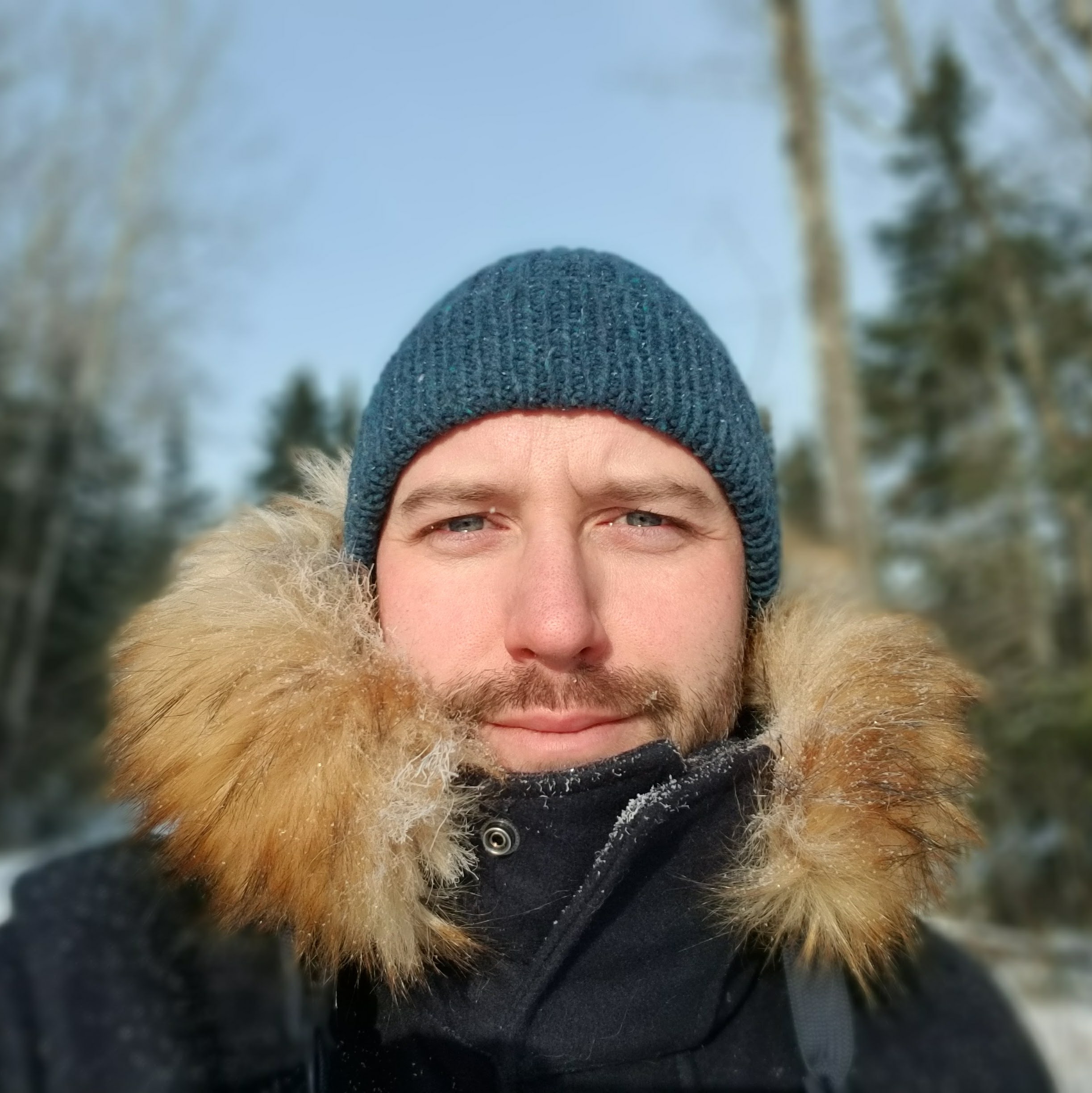 Magnus-Johnston wearing a sock hat and a winter coat with a thick collar, sprinkled with snow, with trees rising behind him.