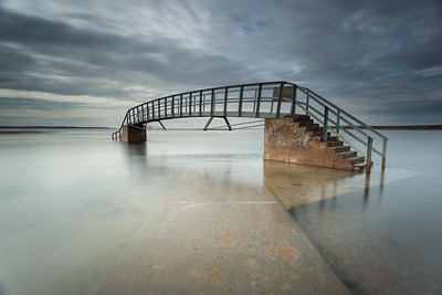 Photograph of a short steel bridge under a gray sky, surrounded on all sides by what seems like a shallow lake