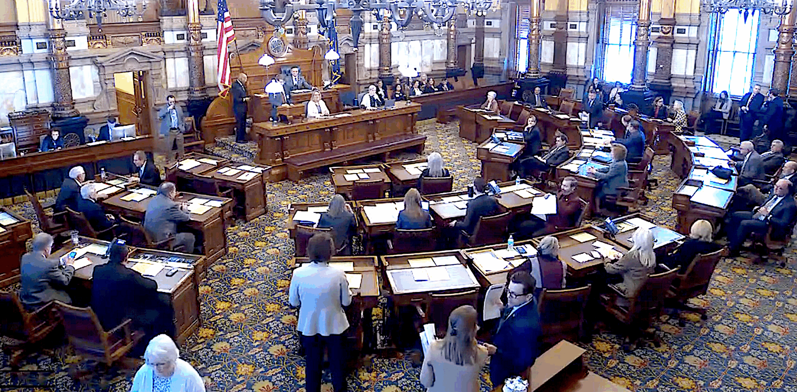 A legislative room with a with desks arranged in an ark around a large desk and podium at the front, with many people in suits sitting at desks and standing around.