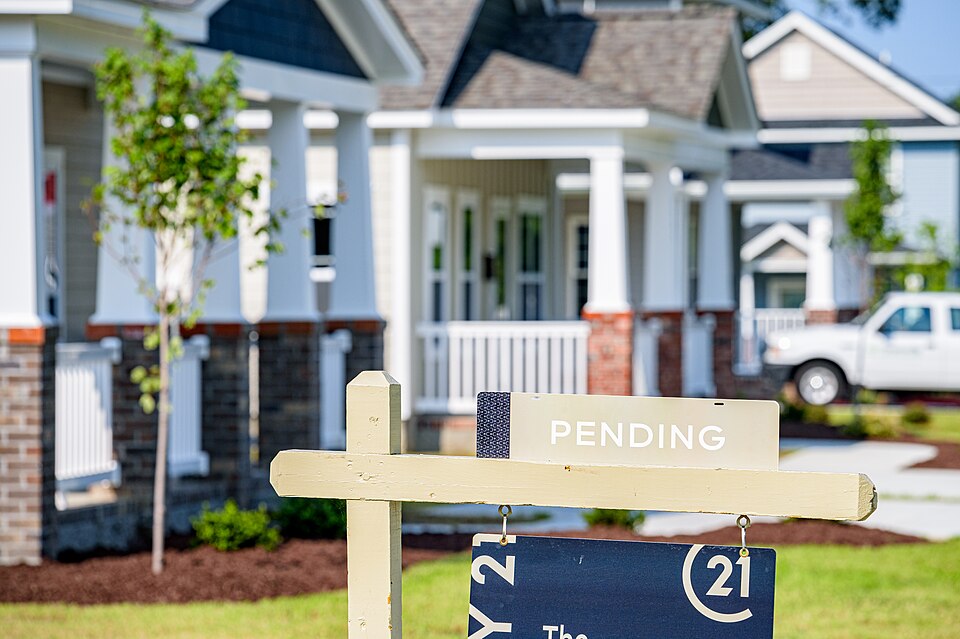 A view looking past a “Pending” home sale sign in front of a row of new homes with fresh paint and landscaping on a sunny day in Greenville, North Carolina.