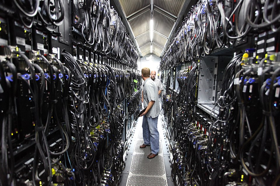 Two employees in a narrow “hot aisle” at a data center walk between tall walls of cords attached to the backs of server racks.