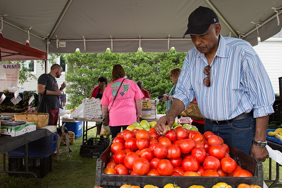 A person wearing a ball cap looks over a crate of red tomatoes as people shop at a table behind him.