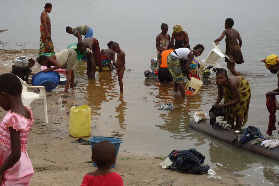 Women and children stand on a shore and in shallow water, many of them bending over to gather water and wash clothes.
