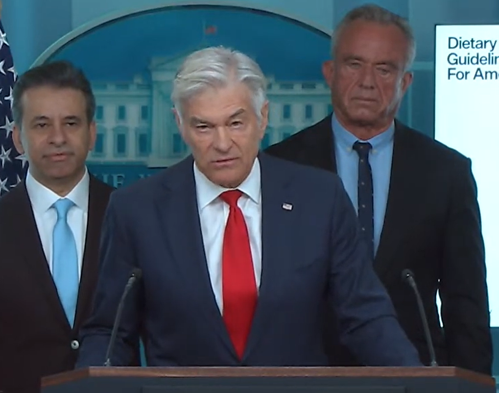 Oz looks into the camera while speaking at a podium, with two men in suits standing behind him.