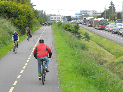 several people bike along a bike path with trees to the left and a traffic-jammed road to the right