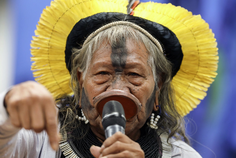 An elderly indigenous man wearing traditional garb and makeup speaks into a microphone and points down at the earth.