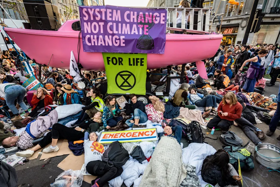 A crowd of people lay and stand in the street around a pink boat with protest banners.