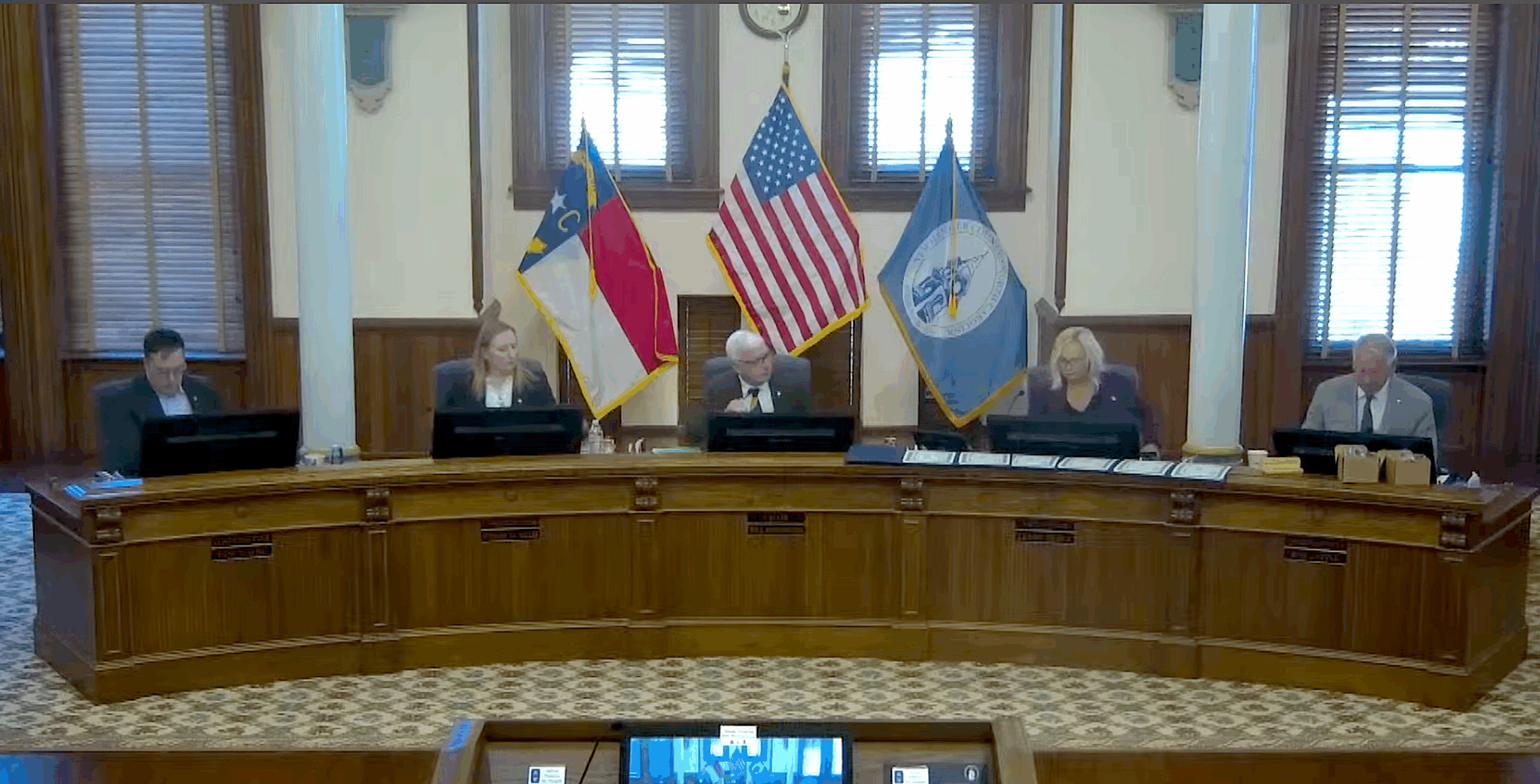 Five people sit behind a podium at the front of a room, with three flags behind them.