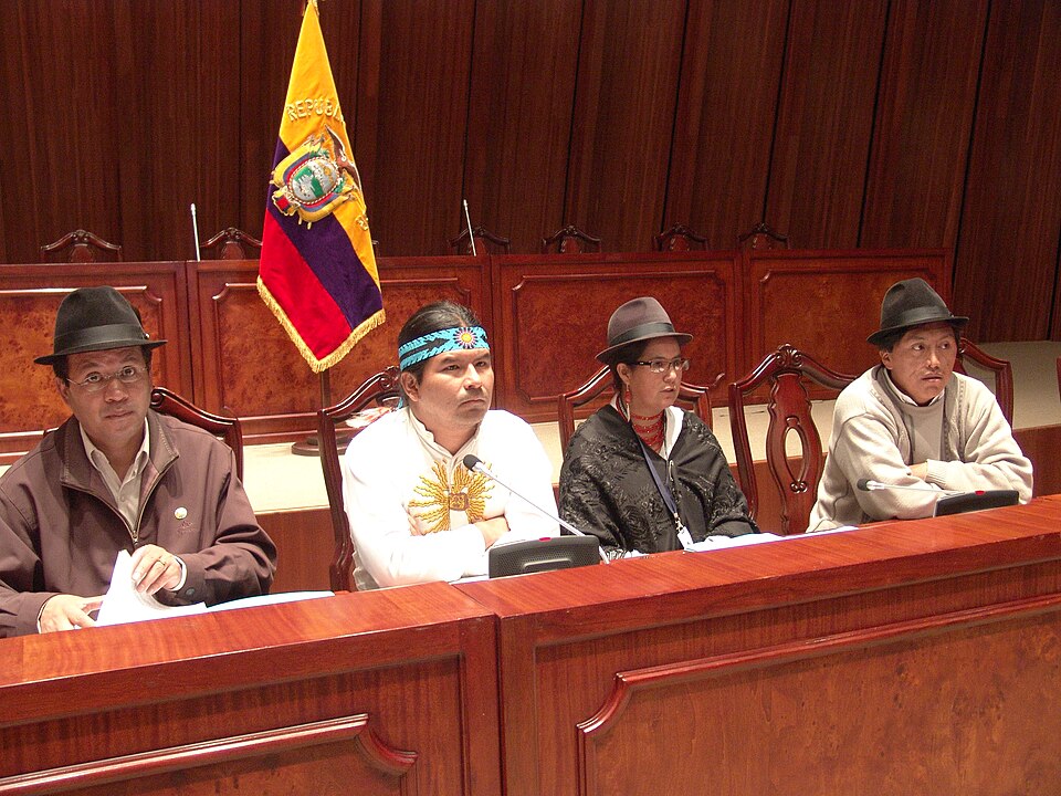 Four people sit in an assembly hall with the Ecuadorian flag behind them. Two of them are wearing indigenous garb.