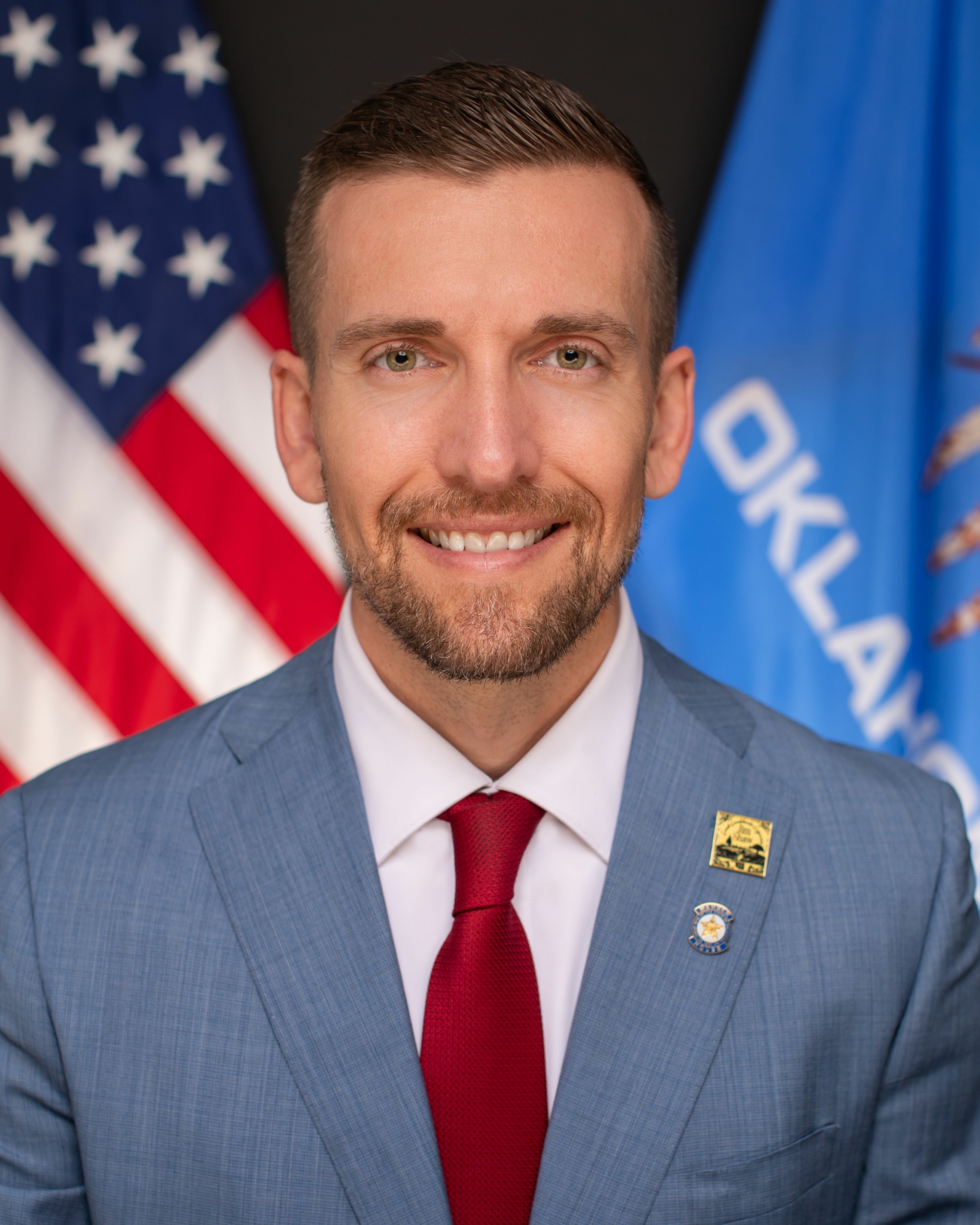 man in a suit and tie smiles in front of U.S. and Oklahoma flags