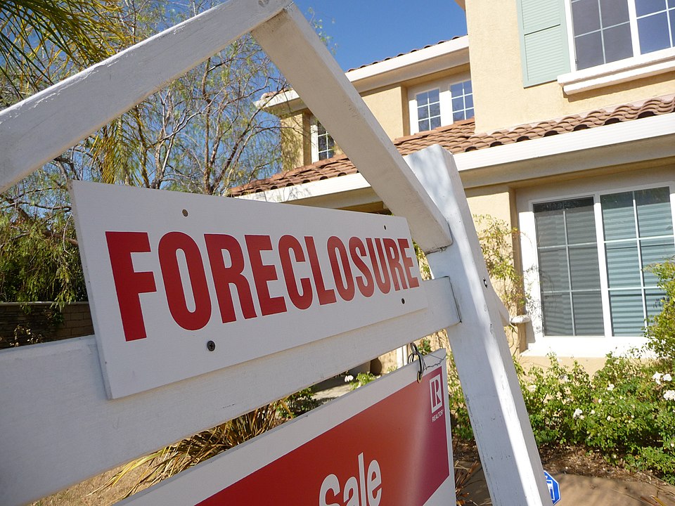 Close up, angled view of a foreclosure sign with bright red text in front of a yellow stucco house with a tile roof.