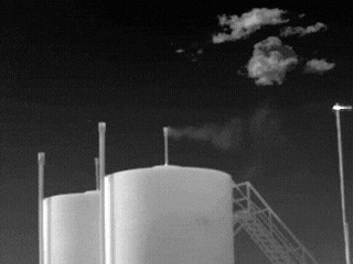 A grayscale thermal image showing infrastructure at an oil and gas processing facility, with a plume of emissions emanating from the top of a storage tank.