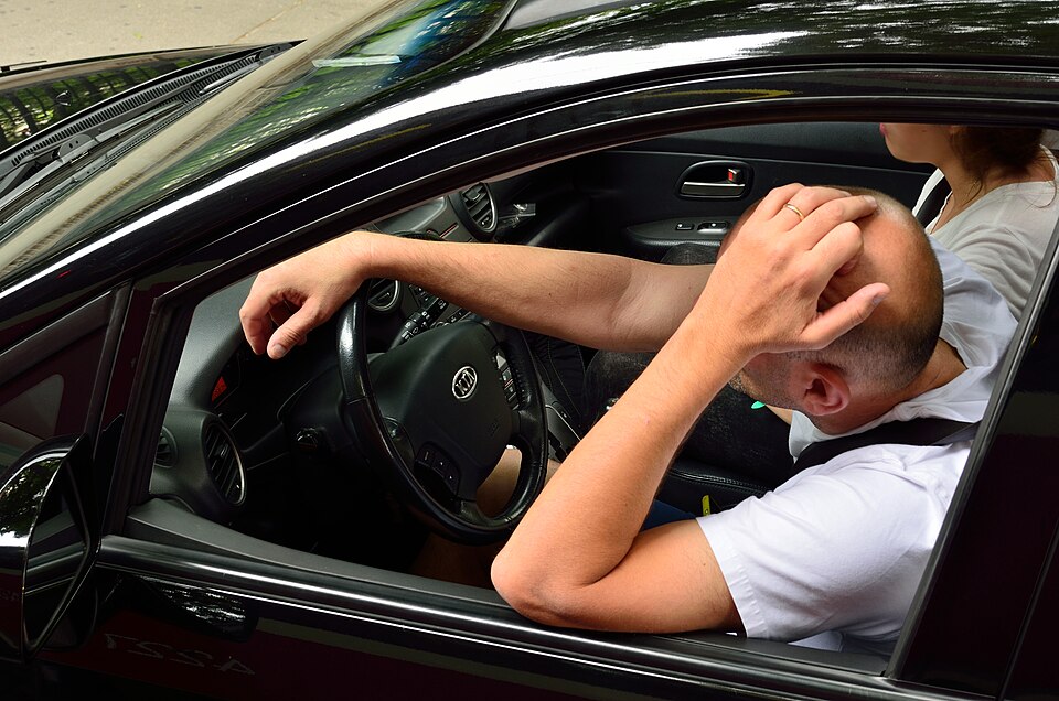 two people sitting in a car, the driver with holding their head in their hand