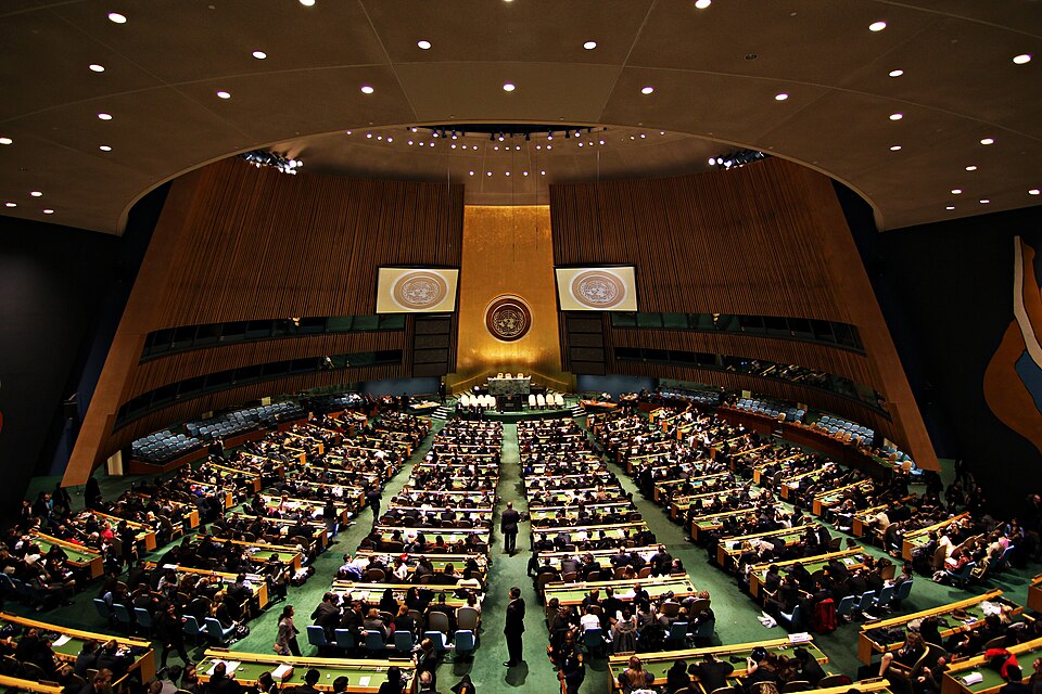 Aerial view of a huge room with the UN symbol on a golden panel at the front and rows of tables full of people.